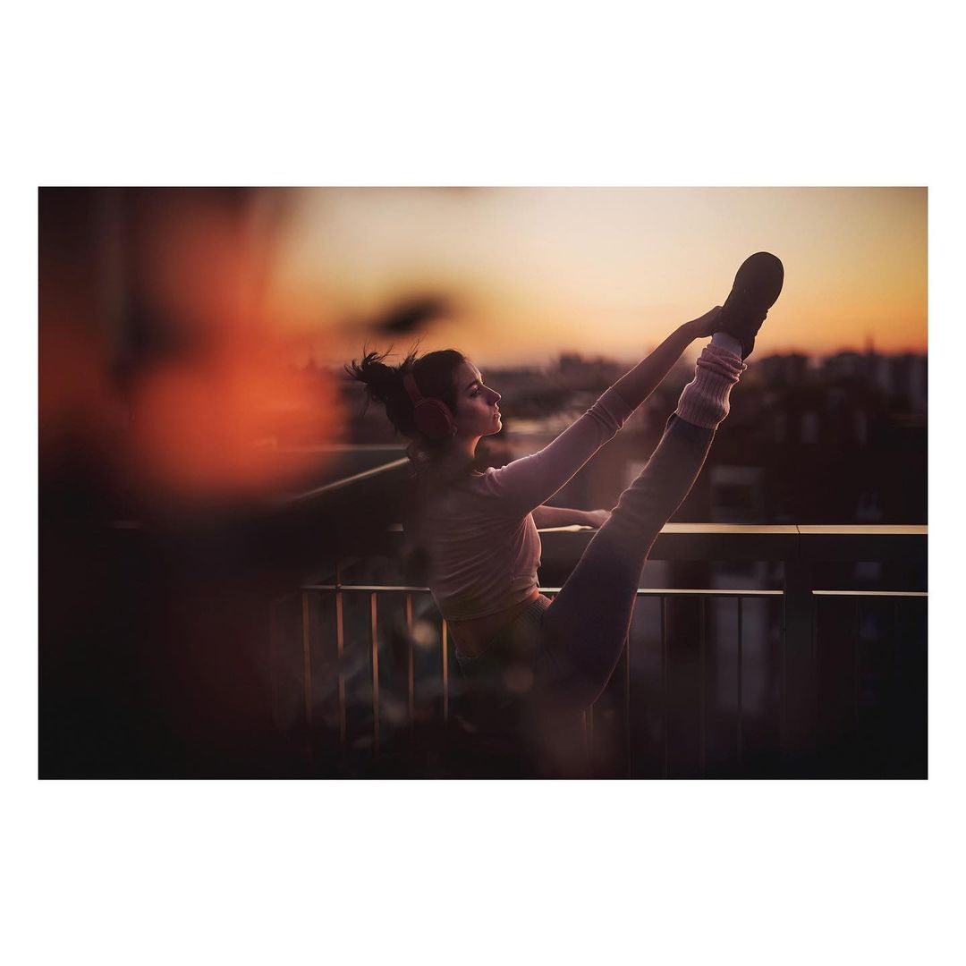 Woman doing yoga on rooftop with sunset in background. She is wearing workout clothes with leg warmers and has her legs up.