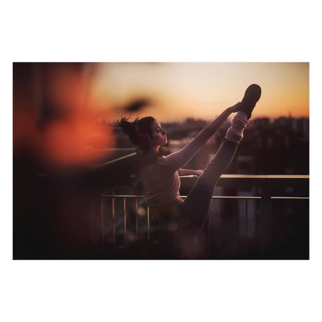 Woman doing yoga on rooftop with sunset in background. She is wearing workout clothes with leg warmers and has her legs up.