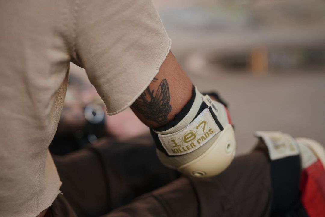 Person with butterfly tattoo wearing cream-colored pads and a beige top poses in a softly-lit scene.