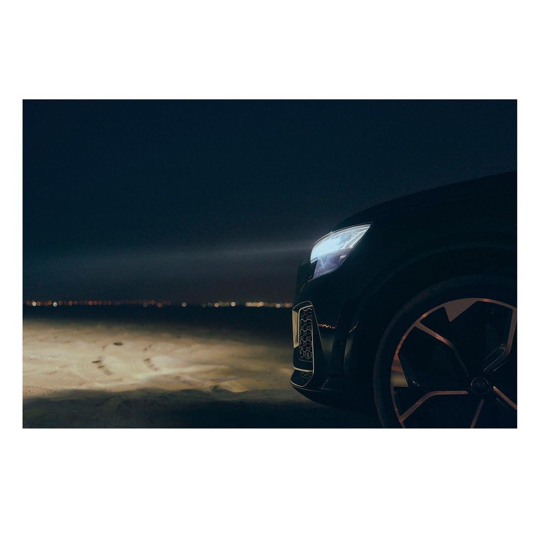 Sleek black car with illuminated headlights, positioned on a dark sandy terrain with city lights faintly visible in the distance.
