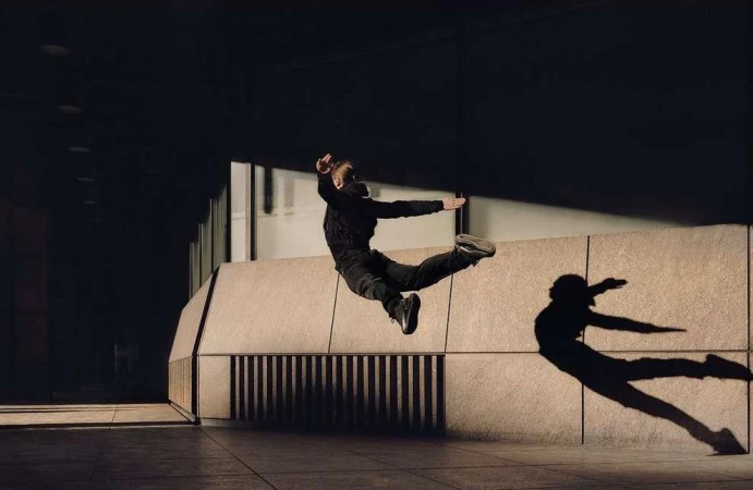 Person in dark clothing mid air leaping horizontally against a light stone wall with a dramatic shadow