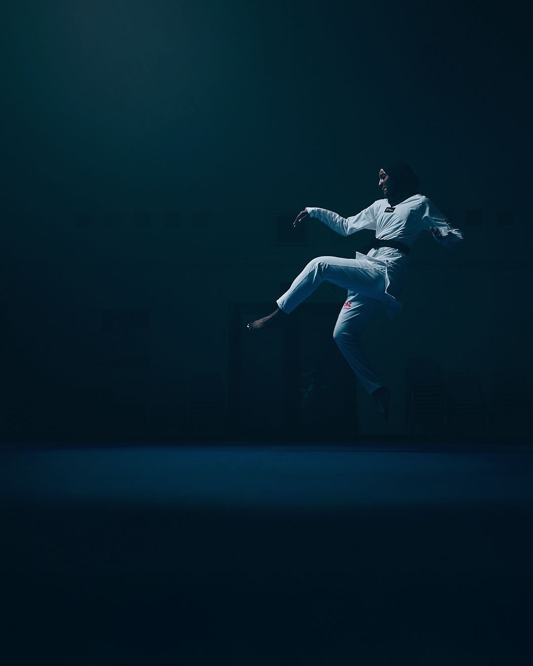 Person in white gi performing a jump kick in a dimly lit indoor martial arts studio with blue lighting. Sport and fitness concept.