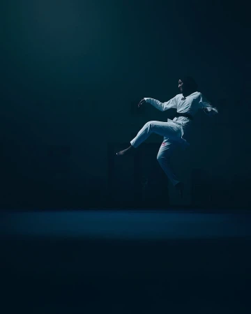 Person in white gi performing a jump kick in a dimly lit indoor martial arts studio with blue lighting. Sport and fitness concept.