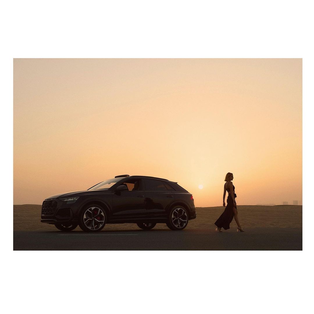 Black SUV with a woman walking along a desert road at sunset. The car is parked on the roadside, and a woman is walking next to it.