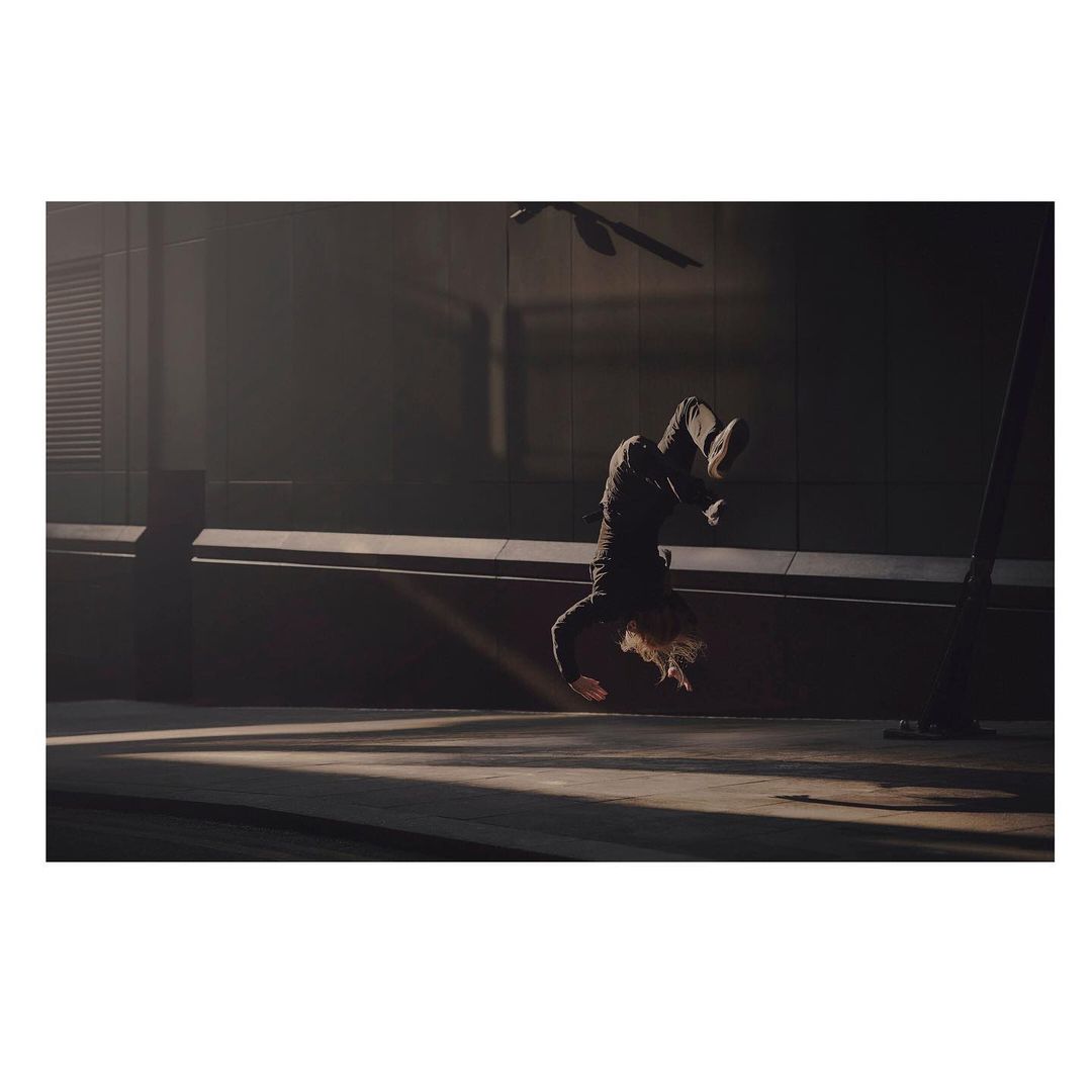 Person in black clothing performing a handstand on a concrete surface with dramatic lighting and architectural backdrop, showcasing fitness and urban style.