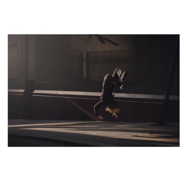 Person in black clothing performing a handstand on a concrete surface with dramatic lighting and architectural backdrop, showcasing fitness and urban style.