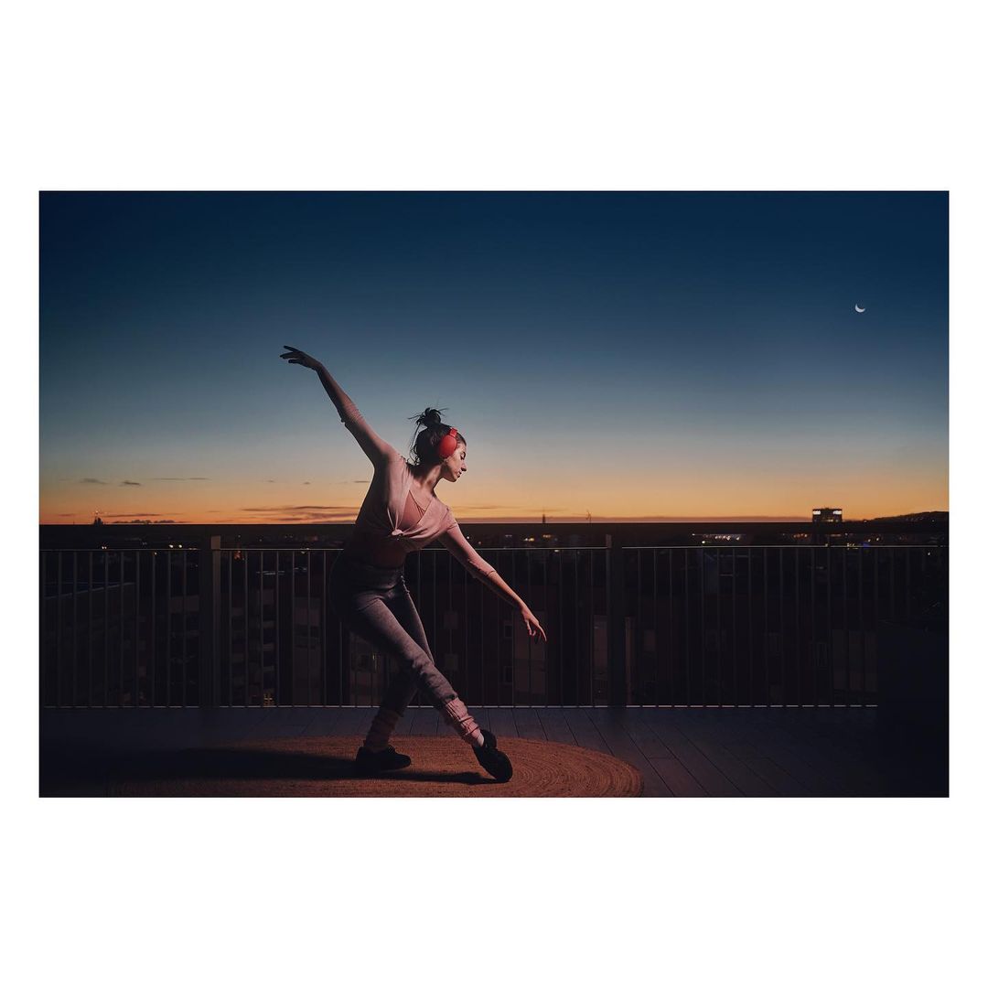 Ballet dancer strikes a pose on a rooftop at dusk. She wears athletic gear, including ballet shoes and leg warmers. A city skyline is visible.