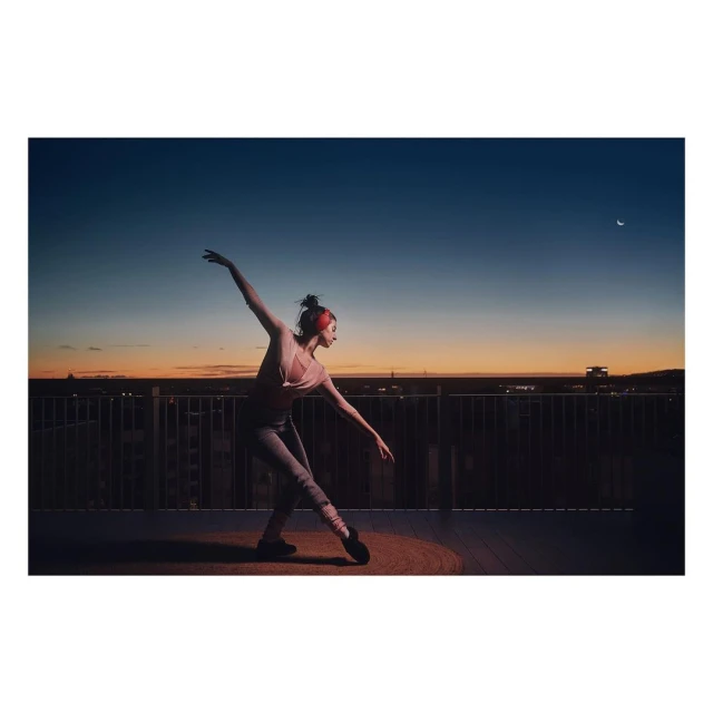 Ballet dancer strikes a pose on a rooftop at dusk. She wears athletic gear, including ballet shoes and leg warmers. A city skyline is visible.