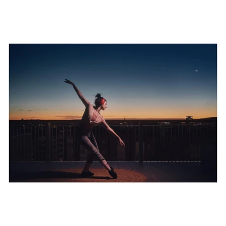 Ballet dancer strikes a pose on a rooftop at dusk. She wears athletic gear, including ballet shoes and leg warmers. A city skyline is visible.