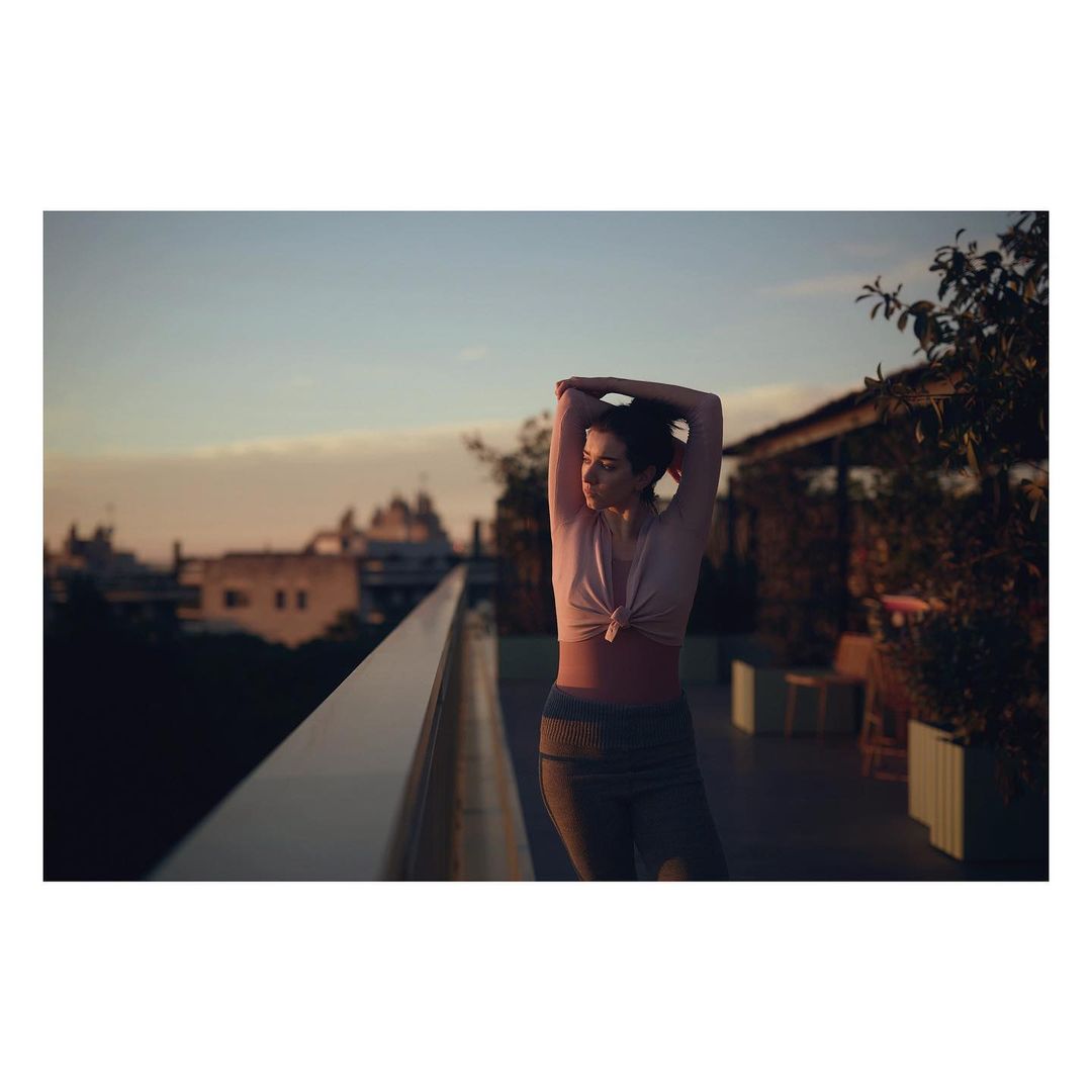 Woman stretches on rooftop at sunrise in athleisure wear with urban background and soft lighting, capturing a peaceful moment.