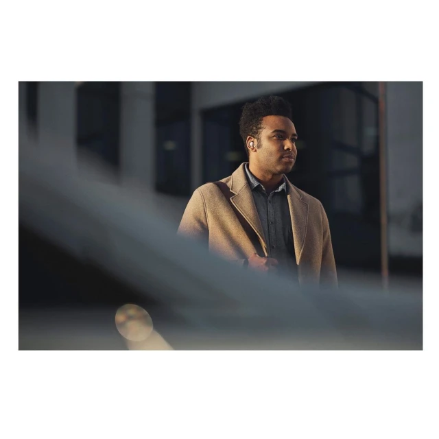An adult male stands outside wearing a tan coat and gray shirt. He is seen from the chest up with buildings in the background.