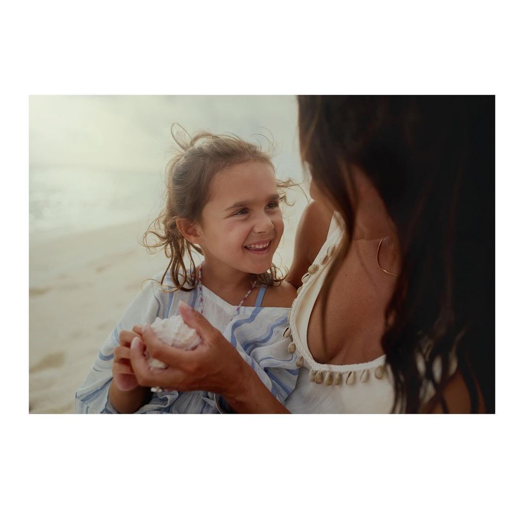 A young girl holds a shell on a beach with an adult female, both smiling; outdoor scene with sand and muted lighting.