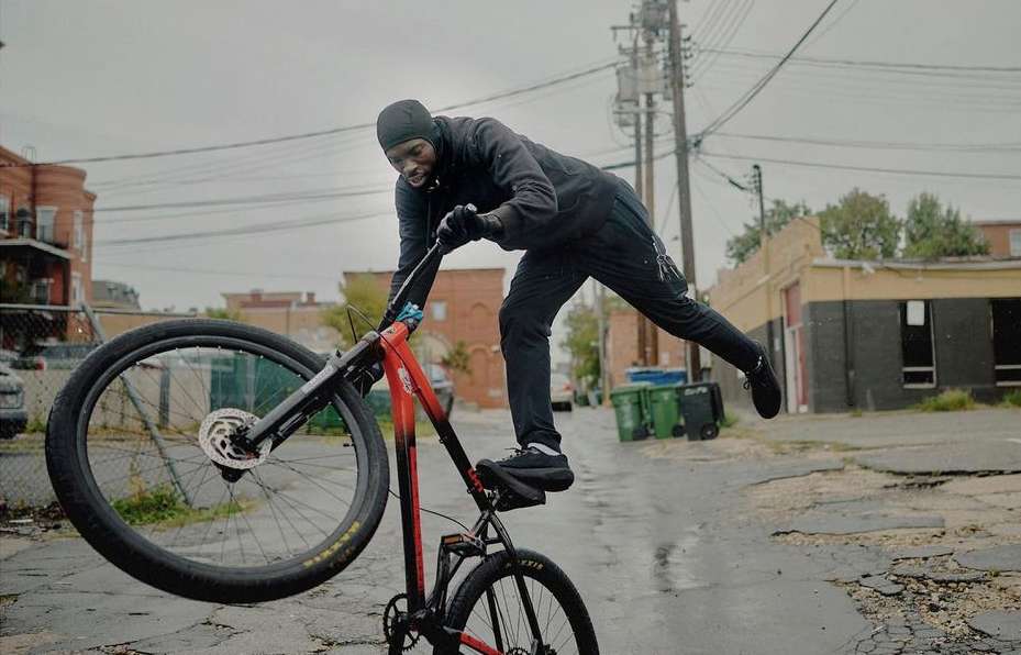 A man performs a stunt on a red and black bicycle on a wet, cracked street in an urban environment under an overcast sky.