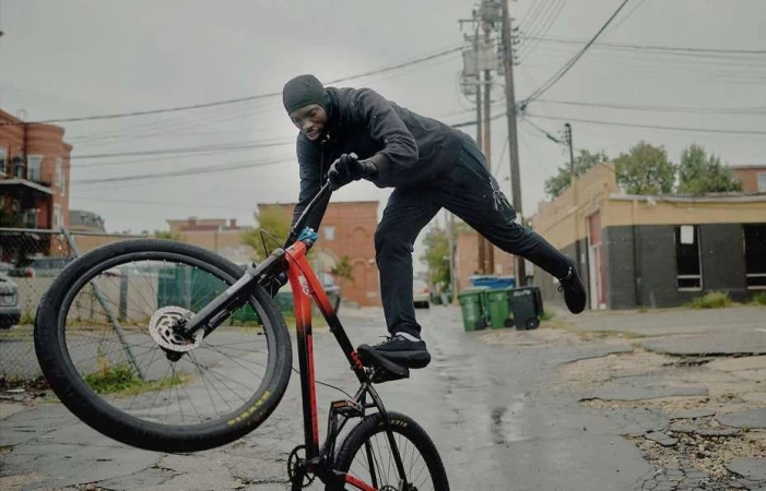 A man performs a stunt on a red and black bicycle on a wet, cracked street in an urban environment under an overcast sky.