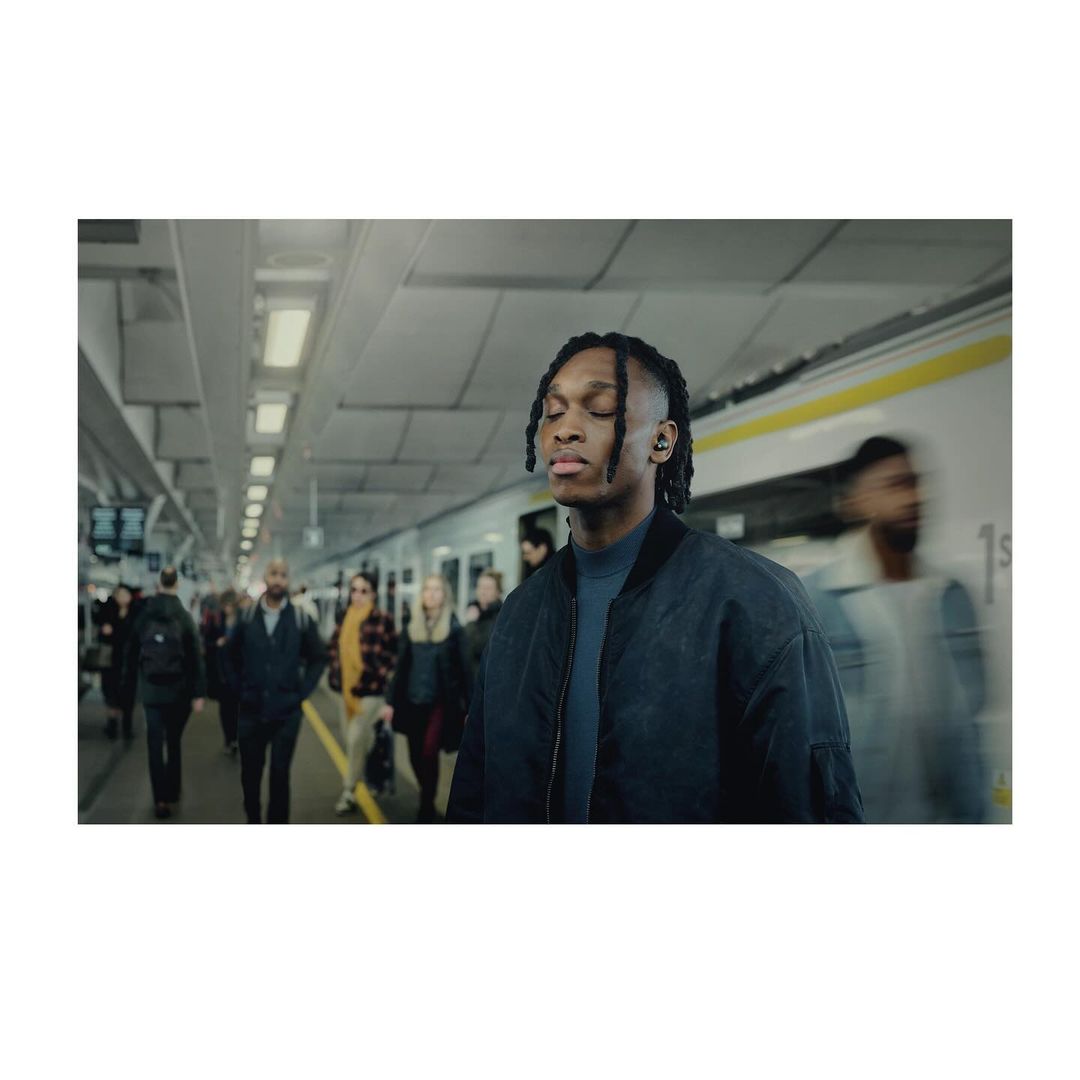 A young Black man with dreadlocks stands with eyes closed on a subway platform, a train blurred in the background with other commuters.