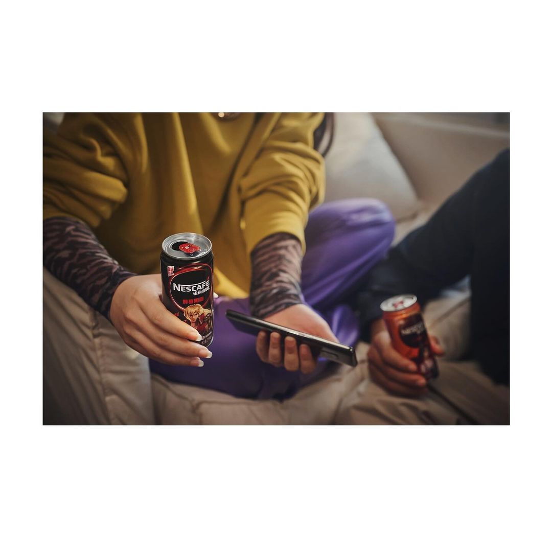Two people on a couch holding cans of Nescafe and a smartphone, low angle, indoor setting, casual clothing, neutral lighting.