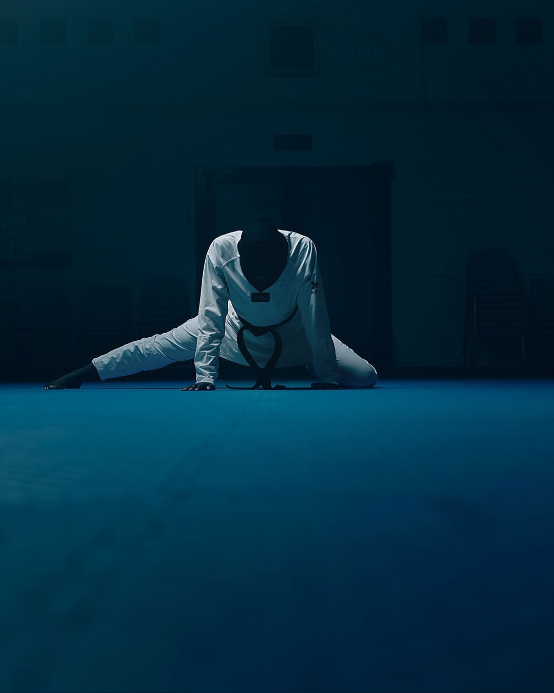 A person in a white uniform does a split on a blue floor in a dimly lit room with a dark background and a closed door.