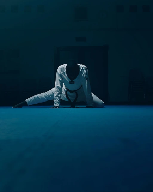 A person in a white uniform does a split on a blue floor in a dimly lit room with a dark background and a closed door.