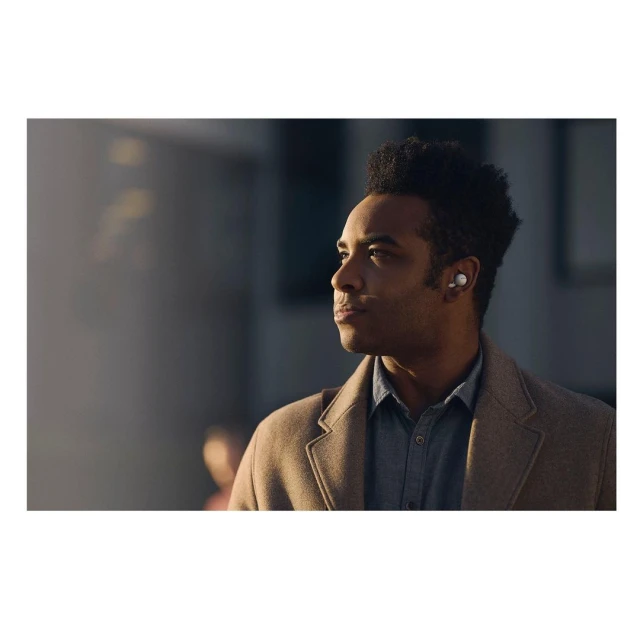 An adult man wearing a coat and wireless earbuds gazes into the distance in an outdoor urban setting with sunlight on his face.