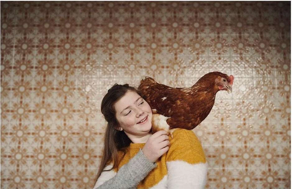 Young woman smiling with a brown hen perched on her shoulder against a patterned tile wall in an indoor setting with soft lighting.