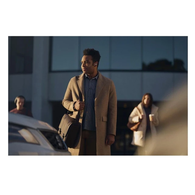 A stylish man with a messenger bag wears wireless earbuds outdoors in an urban setting, with two blurred people and a car in the background.