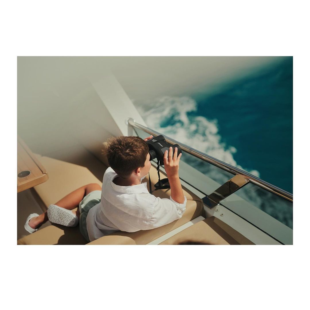 Boy lies on a boat, looking through binoculars at the blue sea and sky, wearing a white shirt and shorts, enjoying the sun.