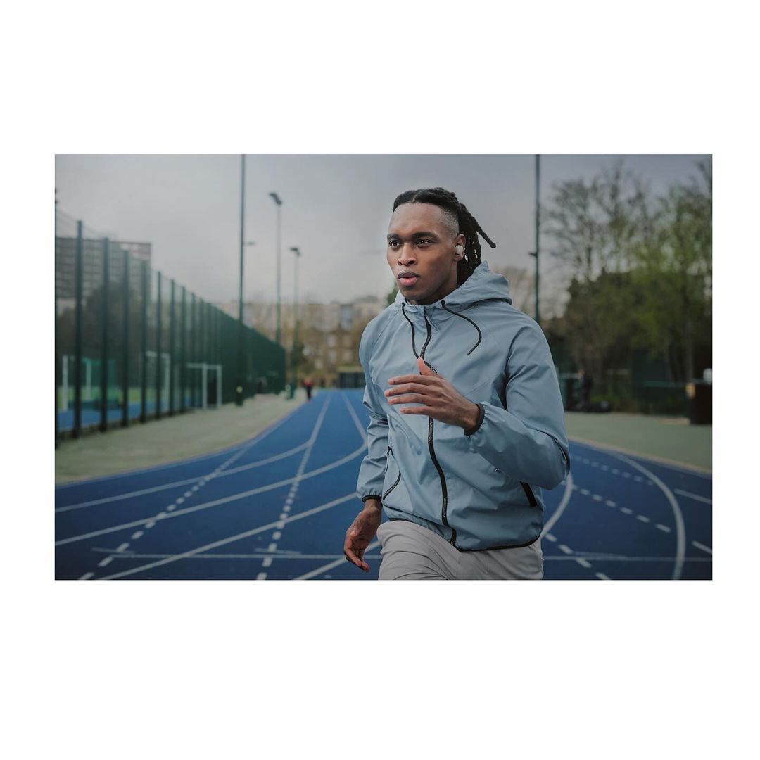 Athletic Black man in a jacket and grey pants jogging on a blue outdoor running track in daylight, focused on fitness and activity.