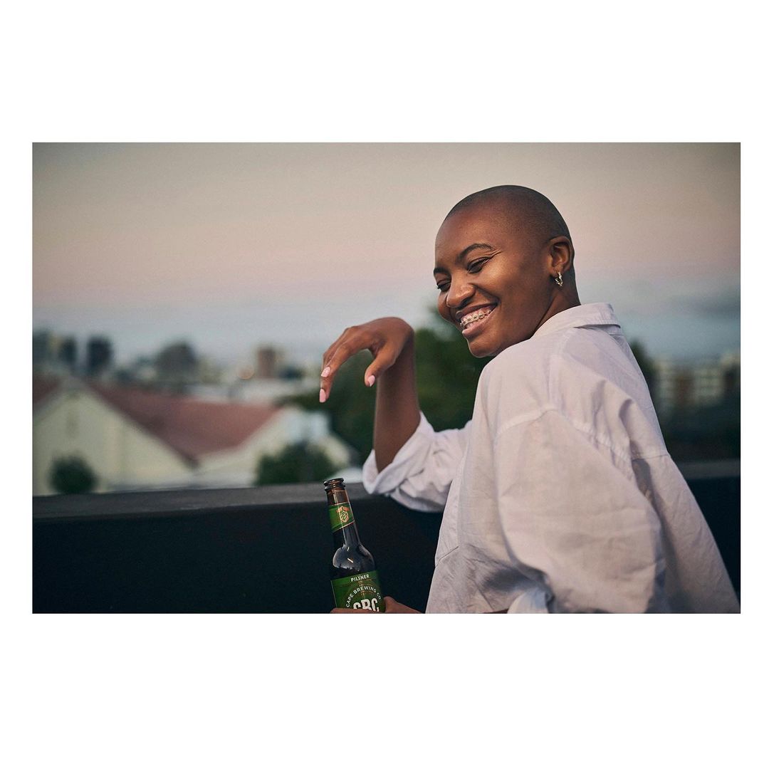 Happy woman in a white shirt smiles while holding a beer on a rooftop at dusk in an urban setting with buildings in the background.