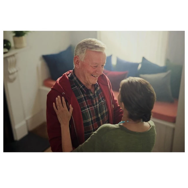 Smiling elderly couple embraces indoors near a window. The man wears a plaid shirt and the woman wears a dark green top.