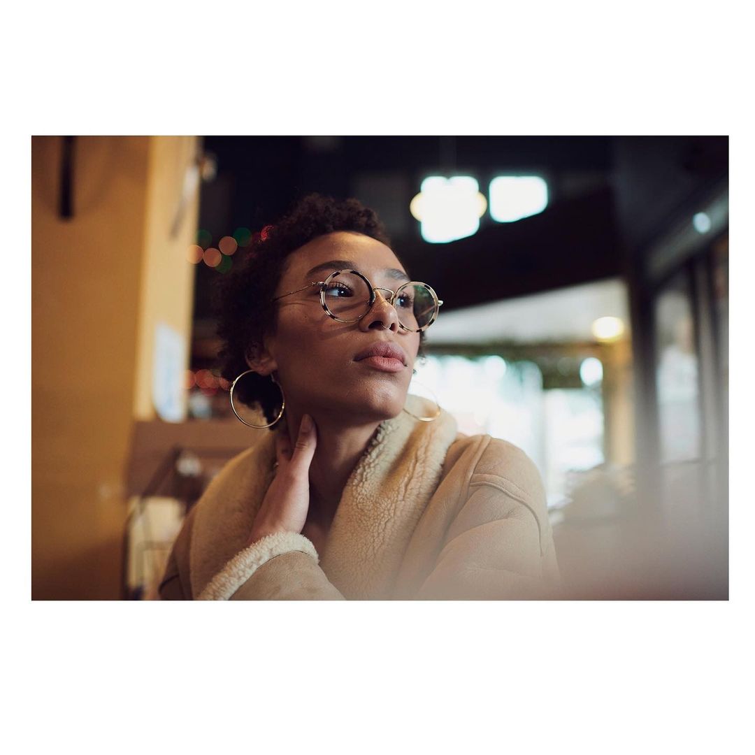 Portrait of a Black woman with curly hair, wearing glasses and a tan jacket, in an indoor setting with bokeh lights in the background.