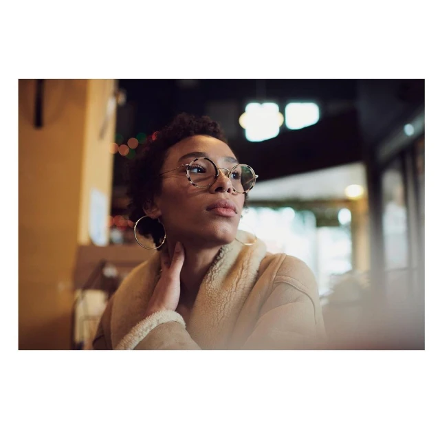 Portrait of a Black woman with curly hair, wearing glasses and a tan jacket, in an indoor setting with bokeh lights in the background.