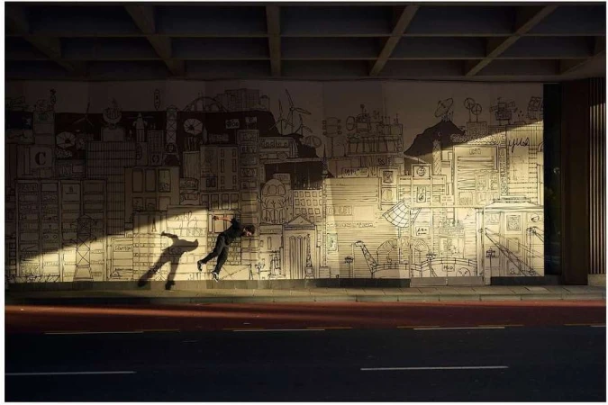 Adult male performing a stunt jump in front of a mural art illustration of a cityscape in an urban environment with visible sunlight.