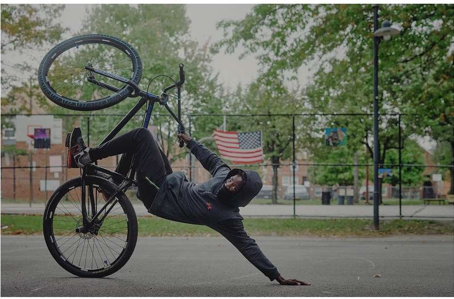 An adult male performs a bicycle trick on an urban court, displaying balance and skill under an overcast sky, with a US flag visible.