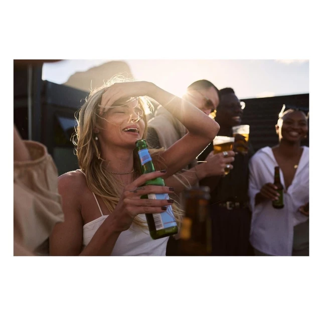 A group of diverse young adults are enjoying an outdoor gathering at sunset, drinking beer and laughing together in a social setting.