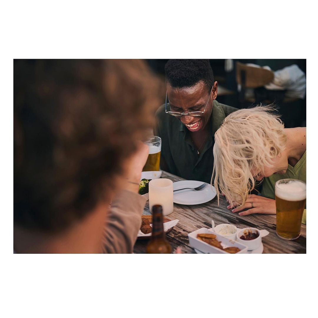 Three individuals laugh together at a wooden table laden with food and beer, creating a warm, convivial atmosphere in an indoor setting.