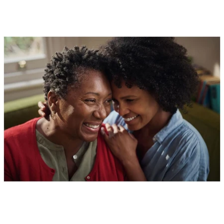 Two African American women share a joyful embrace indoors; one in red, the other in denim, bathed in soft, natural light.