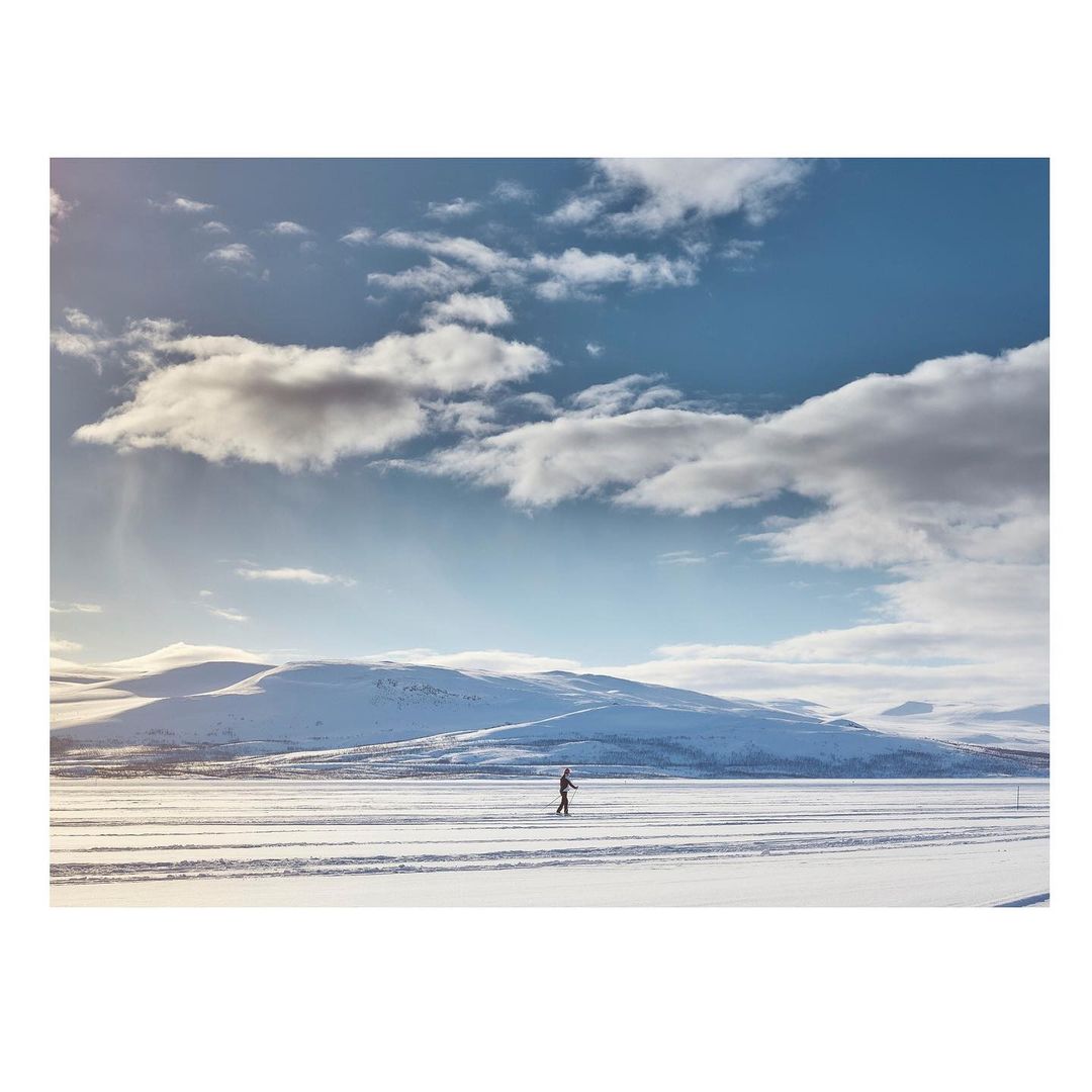 A distant figure cross-country skiing on a snow-covered plain, with snowy mountains in the distance under a blue sky with scattered clouds.