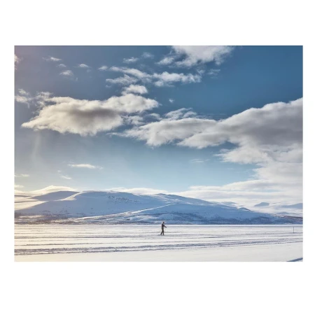A distant figure cross-country skiing on a snow-covered plain, with snowy mountains in the distance under a blue sky with scattered clouds.