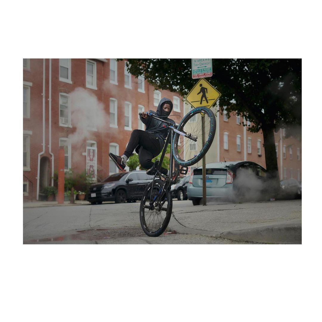 A person on a bicycle does a wheelie on a city street with apartment buildings, cars, and a crosswalk sign in the background.