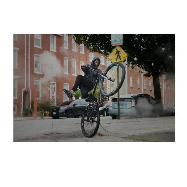 A person on a bicycle does a wheelie on a city street with apartment buildings, cars, and a crosswalk sign in the background.