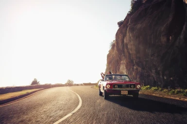 Red convertible car driving along a winding coastal road beneath towering rocky cliffs in bright daylight