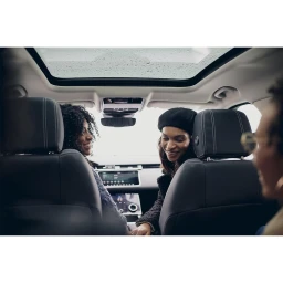 Three people smiling inside a car with a large glass sunroof showing an overcast sky above.