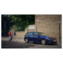 Blue hatchback car parked on a London street with two people walking in the background near a brick wall.