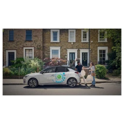 Man and woman loading white car with shopping bags parked on a London street in front of brick residential buildings.