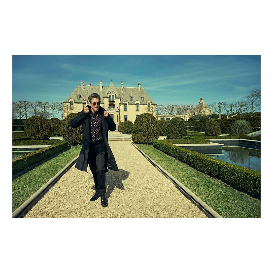 Mature man in a black coat walks on a gravel path toward a large estate, set against a blue sky in an outdoor setting.