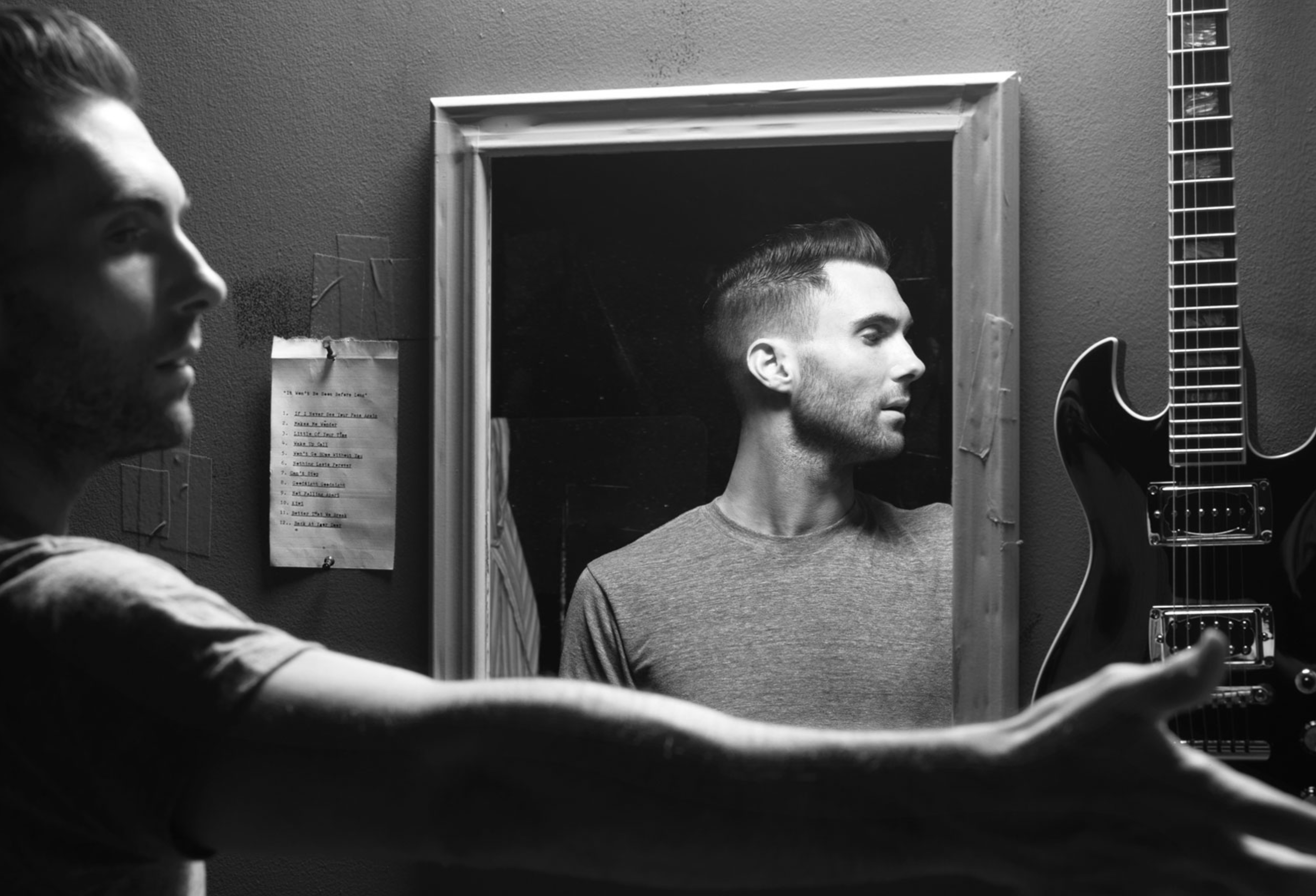 Black and white portrait of a man reflected in a mirror, beside a guitar and a handwritten note on the wall. Studio setting.