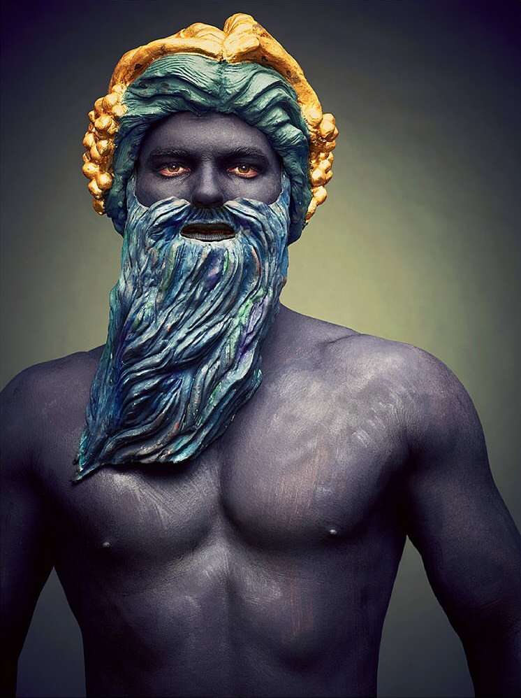 Artistic portrait of a dark-skinned man with a blue beard and hair, wearing a gold headdress against a soft gray backdrop.