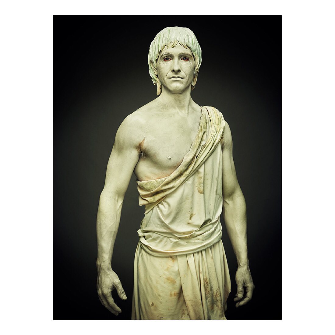 Man with body paint posing in draped fabric against a dark background. Studio shot, portrait of an adult male painted as a statue.
