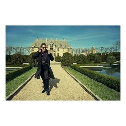 Man in dark coat and sunglasses walking down a gravel path towards a large estate house.