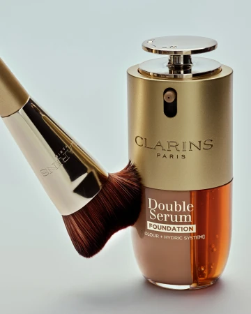 Clarins Double Serum Foundation bottle with a makeup brush resting against it on a grey studio background.